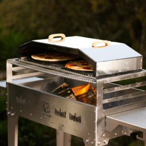 Stainless steel Double Pizza Dome on a wood-fired outdoor grill, baking two pizzas to perfection in an open-air setting.